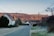 A residential street in a rural area with houses lining both sides. The road is quiet and leads towards rolling hills in the background, illuminated by the soft glow of a setting or rising sun. Telephone poles and wires stretch across the sky, and trees and bushes are seen on either side of the street.