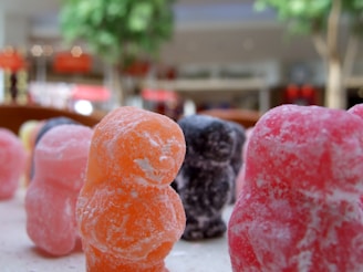 Close-up view of colorful, sugar-coated gummy candies shaped like small bears, with blurred trees and a mall interior in the background.