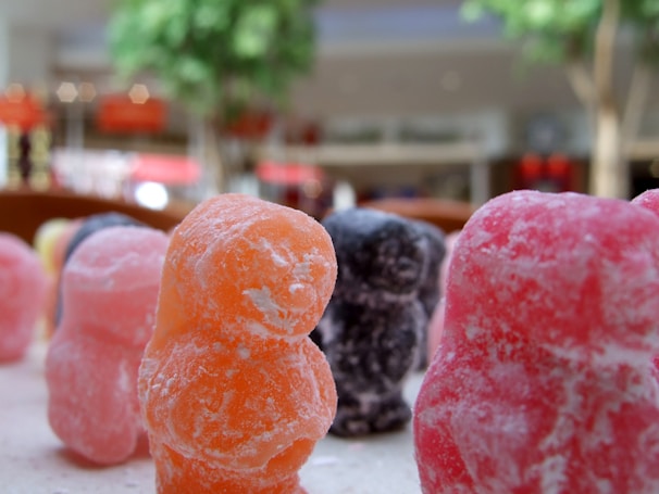 Close-up view of colorful, sugar-coated gummy candies shaped like small bears, with blurred trees and a mall interior in the background.