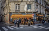 A corner bakery with a blue and yellow facade, displaying the words 'Boulanger' and 'Pâtissier'. People stand on the sidewalk near a pedestrian crossing, some engaged in conversation. The shop window reveals a warm, inviting interior with several patrons seated inside.