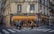 A corner bakery with a blue and yellow facade, displaying the words 'Boulanger' and 'Pâtissier'. People stand on the sidewalk near a pedestrian crossing, some engaged in conversation. The shop window reveals a warm, inviting interior with several patrons seated inside.