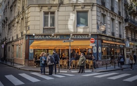 A corner bakery with a blue and yellow facade, displaying the words 'Boulanger' and 'P&acirc;tissier'. People stand on the sidewalk near a pedestrian crossing, some engaged in conversation. The shop window reveals a warm, inviting interior with several patrons seated inside.