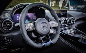 A luxury car interior featuring a sleek design with a focus on the steering wheel. The dashboard includes modern digital displays and metallic accents, enhancing the overall elegance and sophistication. The ambient lighting and polished surfaces reflect a high level of craftsmanship.
