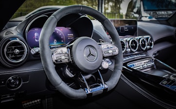 A luxury car interior featuring a sleek design with a focus on the steering wheel. The dashboard includes modern digital displays and metallic accents, enhancing the overall elegance and sophistication. The ambient lighting and polished surfaces reflect a high level of craftsmanship.