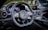 A luxury car interior featuring a sleek design with a focus on the steering wheel. The dashboard includes modern digital displays and metallic accents, enhancing the overall elegance and sophistication. The ambient lighting and polished surfaces reflect a high level of craftsmanship.