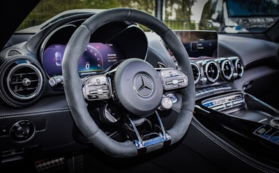 A luxury car interior featuring a sleek design with a focus on the steering wheel. The dashboard includes modern digital displays and metallic accents, enhancing the overall elegance and sophistication. The ambient lighting and polished surfaces reflect a high level of craftsmanship.