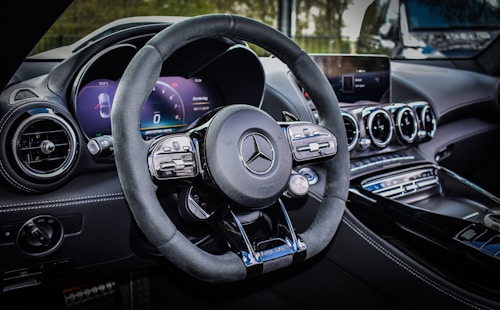 A luxury car interior featuring a sleek design with a focus on the steering wheel. The dashboard includes modern digital displays and metallic accents, enhancing the overall elegance and sophistication. The ambient lighting and polished surfaces reflect a high level of craftsmanship.