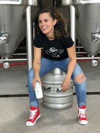 A person is sitting on a metal beer keg, smiling and holding a can. They are wearing a black t-shirt, ripped blue jeans, and bright red high-top sneakers. The background shows large stainless steel fermentation tanks in a brewery setting.