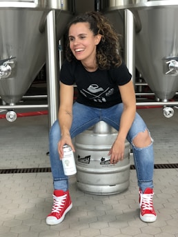 A person is sitting on a metal beer keg, smiling and holding a can. They are wearing a black t-shirt, ripped blue jeans, and bright red high-top sneakers. The background shows large stainless steel fermentation tanks in a brewery setting.