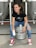 A person is sitting on a metal beer keg, smiling and holding a can. They are wearing a black t-shirt, ripped blue jeans, and bright red high-top sneakers. The background shows large stainless steel fermentation tanks in a brewery setting.