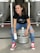 A person is sitting on a metal beer keg, smiling and holding a can. They are wearing a black t-shirt, ripped blue jeans, and bright red high-top sneakers. The background shows large stainless steel fermentation tanks in a brewery setting.