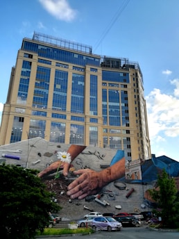 A large mural painted on the side of a building depicts two hands tending to the soil, one of which is holding a daisy. The mural includes various tools and objects scattered around the hands. The building itself is a tall, modern structure with many windows, and the sky is clear and blue.