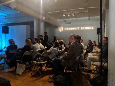 A room with several people seated in folding chairs, attentively facing a presentation area. The environment is professional, likely a seminar or workshop, with 'Product School' illuminated on a wall. Some attendees are taking notes, and a few people are standing, possibly facilitating the event.