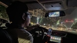 A person is driving a car at night on a city street. The interior of the car shows illuminated dashboard controls and a smartphone mounted on the dashboard displaying GPS navigation. The cityscape outside is dimly lit with streetlights and blurred building lights visible through the windshield.