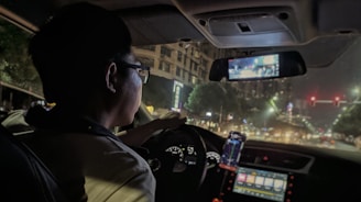 A person is driving a car at night on a city street. The interior of the car shows illuminated dashboard controls and a smartphone mounted on the dashboard displaying GPS navigation. The cityscape outside is dimly lit with streetlights and blurred building lights visible through the windshield.