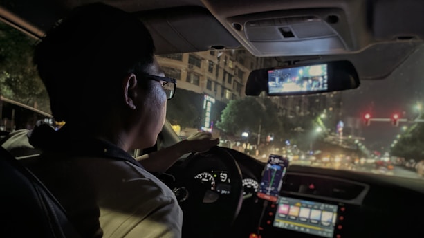 A driver using a smartphone app inside a car at dusk with city lights in the background