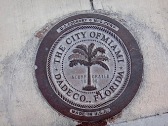 A round metal manhole cover embedded in concrete, featuring the text 'The City of Miami, Dade Co., Florida' surrounding an image of a palm tree. The cover is marked with 'Incorporated 1896' and 'Made in U.S.A.', manufactured by U.S. Foundry & Mfg. Corp.