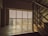 A large window with frosted glass panels allowing diffused light to enter, casting a warm glow on a concrete floor. Metal railings of a staircase are visible on the right, indicating the interior setting of a building.