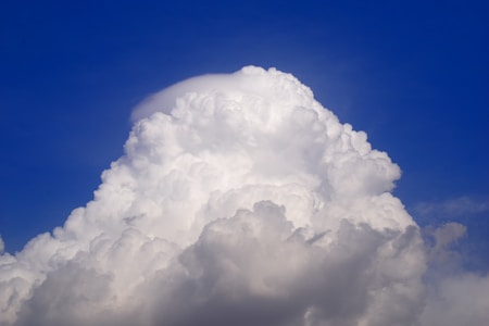 A large, billowing white cloud rises majestically against a clear blue sky. The cloud has a towering, fluffy appearance, with bright sunlight highlighting the top edges, creating subtle shadows within the cloud's texture.