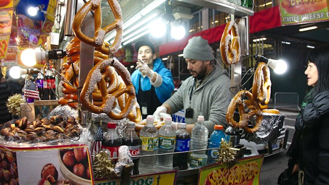 A bustling street vendor stall with large pretzels prominently displayed. Two vendors are serving customers, with one wearing a grey beanie and another gesturing in the background. Various drinks in plastic bottles, along with other snacks, are visibly arranged on the counter. Bright lights illuminate the scene, and small American flags add decorative touches.