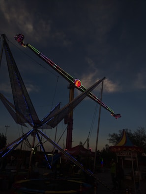 A nighttime amusement park scene with a tall ride featuring neon lights ascending towards the sky. The foreground includes a colorful conical tent and a ticket booth labeled 'Taquilla'. The sky is dark with some clouds visible.
