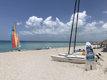 A sandy beach with several catamarans resting on the shore. A colorful sailboat with vibrant hues of orange, yellow, and blue is prominently visible. The ocean is calm and a mix of green and blue under a partly cloudy sky. On the right, a thatched hut with beach chairs can be seen.