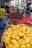 A vibrant street market scene with a variety of fruits on display. Foreground is filled with a large pile of bright yellow mangoes, while the background shows red lychees and cherries. People walk along the street and browse through the stalls. A colorful umbrella adds a lively touch to the market atmosphere.