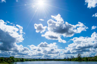 Sunlight streaming through blue skies above a calm lake surrounded by trees.