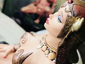 A close-up view of a detailed sculpture or figurine depicting a person with ornate armor and accessories. The figure has red lips, blue eye makeup, and reddish-brown headgear with intricate designs. The surface of the sculpture has a smooth and polished finish, showcasing expert craftsmanship.