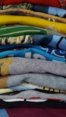 A stack of colorful folded t-shirts with various designs and patterns. The fabrics showcase different textures, colors, and graphic prints including text and images.