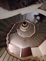 A spiral staircase viewed from above, creating a dizzying, inward spiral pattern that draws the eye to the center. The stairs are surrounded by a muted color scheme, with hints of concrete and metal textures. The blurred figure of a person is visible as they navigate the stairs, suggesting motion.