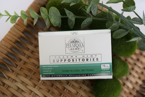 A product labeled 'Pharma Hemp Suppositories' is placed on a woven surface. The packaging details ingredients such as hemp cannabis extract, cocoa butter, coconut oil, and frankincense oil. Green foliage surrounds the product, enhancing a natural aesthetic.