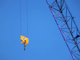 A bright yellow crane hook hanging from cables against a clear blue sky. Part of a crane's metal arm is visible diagonally in the frame, showing a lattice structure.