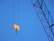 A bright yellow crane hook hanging from cables against a clear blue sky. Part of a crane's metal arm is visible diagonally in the frame, showing a lattice structure.
