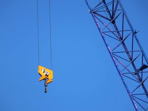 A bright yellow crane hook hanging from cables against a clear blue sky. Part of a crane's metal arm is visible diagonally in the frame, showing a lattice structure.