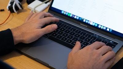 Hands are typing on a MacBook Pro laptop with a visible keyboard and touchpad. The screen displays a website or document with lists. The laptop is placed on a wooden desk alongside an external device connected via cables.