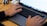 Hands are typing on a MacBook Pro laptop with a visible keyboard and touchpad. The screen displays a website or document with lists. The laptop is placed on a wooden desk alongside an external device connected via cables.