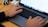 Hands are typing on a MacBook Pro laptop with a visible keyboard and touchpad. The screen displays a website or document with lists. The laptop is placed on a wooden desk alongside an external device connected via cables.