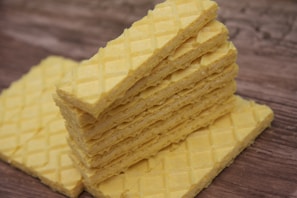 A stack of rectangular, yellow wafer cookies is placed on a wooden surface. The wafers have a crisscross pattern on the top, giving them a textured appearance.