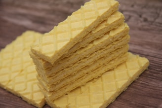 A stack of rectangular, yellow wafer cookies is placed on a wooden surface. The wafers have a crisscross pattern on the top, giving them a textured appearance.