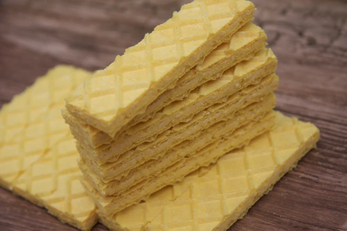 A stack of rectangular, yellow wafer cookies is placed on a wooden surface. The wafers have a crisscross pattern on the top, giving them a textured appearance.