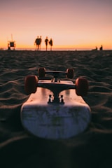A skateboard is positioned on the sand, facing towards a sunset. In the distance, a group of four people stand together, silhouetted against the sky with hues of orange and pink. The scene conveys a peaceful and relaxed beach atmosphere.