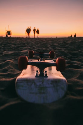 A skateboard is positioned on the sand, facing towards a sunset. In the distance, a group of four people stand together, silhouetted against the sky with hues of orange and pink. The scene conveys a peaceful and relaxed beach atmosphere.