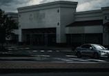 A dark, overcast scene of an empty storefront with a faded sign above. A silver sedan is parked near the entrance. The atmosphere appears gloomy, with cloudy skies in the background and shadows cast on the sidewalk.