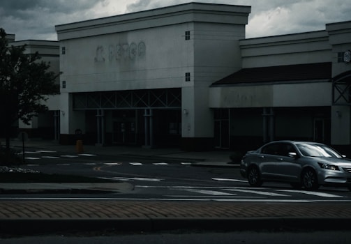 A dark, overcast scene of an empty storefront with a faded sign above. A silver sedan is parked near the entrance. The atmosphere appears gloomy, with cloudy skies in the background and shadows cast on the sidewalk.