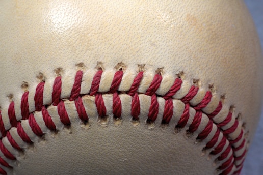 A close-up view of a baseball focusing on the stitching detail. The red stitches are tightly woven into the cream-colored leather surface, showing texture and wear typical of a used baseball.