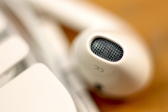A close-up view of a white earbud with a visible mesh speaker and an 'R' marking, resting on a surface that appears to partially show a keyboard key.
