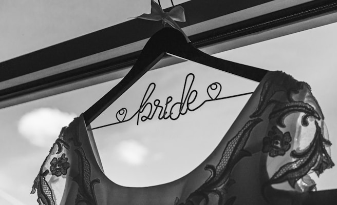 A black and white photograph features a decorative hanger with the word 'bride' elegantly written in wire. A wedding dress with intricate lace patterns and floral embroidery is draped over the hanger. The background shows part of a window with soft, blurred clouds visible outside.