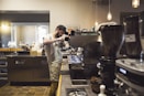 A barista is preparing a coffee drink using an espresso machine in a cozy coffee shop. The shop has a warm and inviting atmosphere with dim lighting and various coffee equipment visible around. Shelves with cups and a counter filled with items like a card reader, coffee grinder, and takeaway cups can be seen.