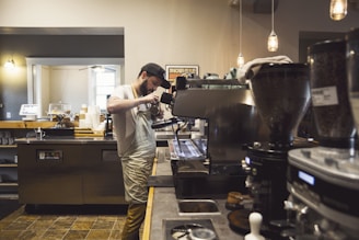 A barista is preparing a coffee drink using an espresso machine in a cozy coffee shop. The shop has a warm and inviting atmosphere with dim lighting and various coffee equipment visible around. Shelves with cups and a counter filled with items like a card reader, coffee grinder, and takeaway cups can be seen.